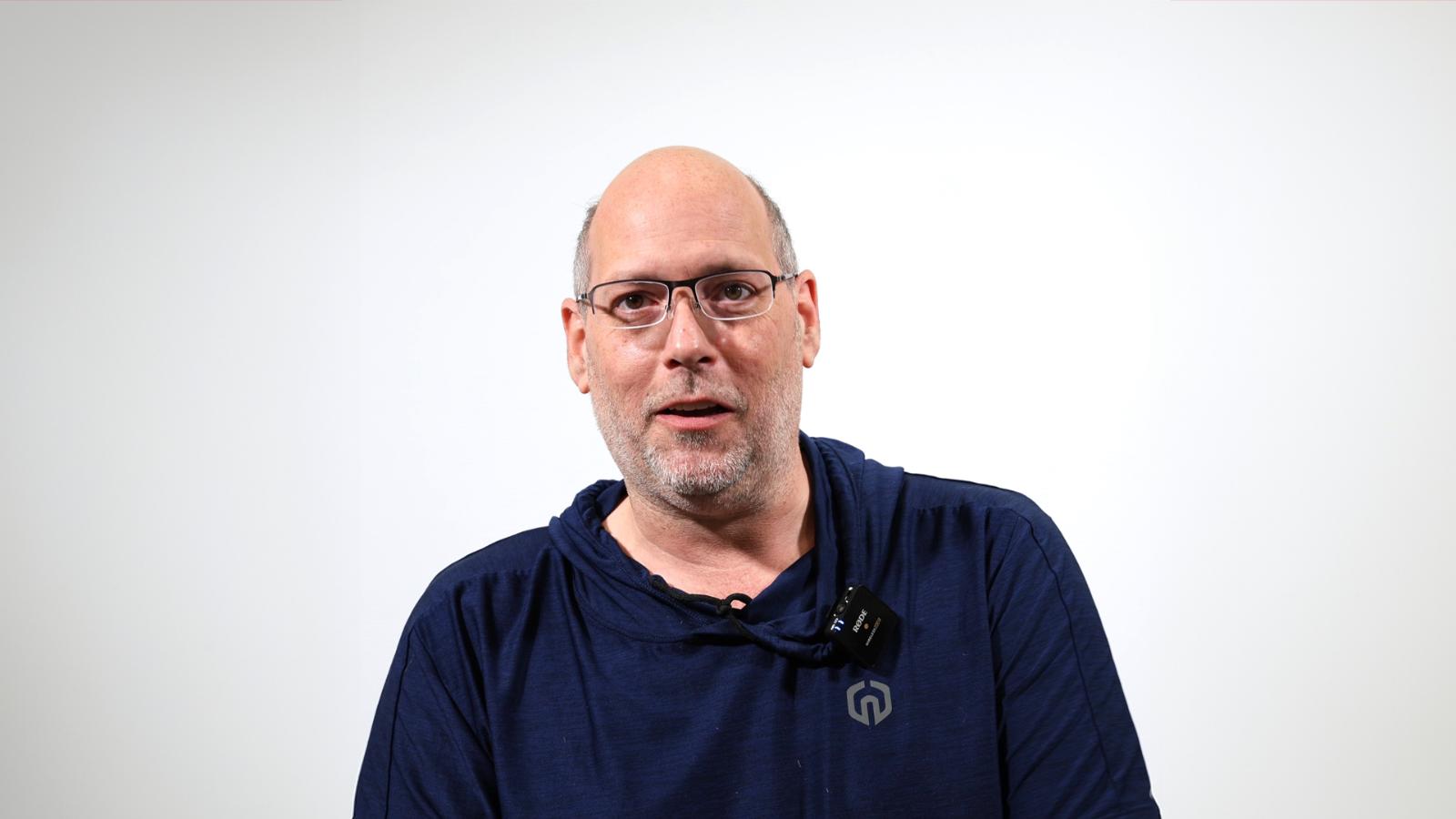 A man with glasses, a short beard, and a bald head wearing a navy blue shirt, facing the camera with a neutral expression against a plain white background.