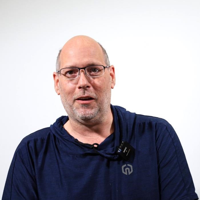 A bald man with glasses and a short beard is wearing a dark blue hoodie with a microphone clipped to it. He is standing against a white background.