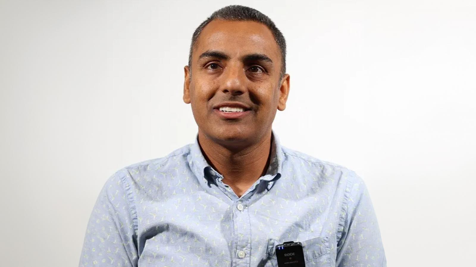 A man wearing a light blue button-up shirt with a small device clipped to it smiles while looking slightly off to the side against a plain white background.