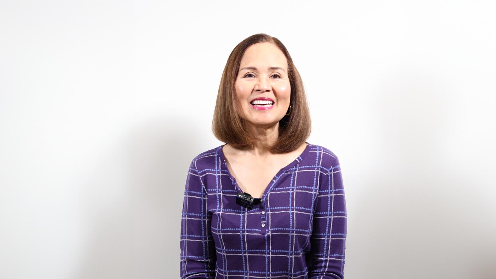 A woman with shoulder-length brown hair, wearing a purple plaid shirt, smiles against a plain white background.