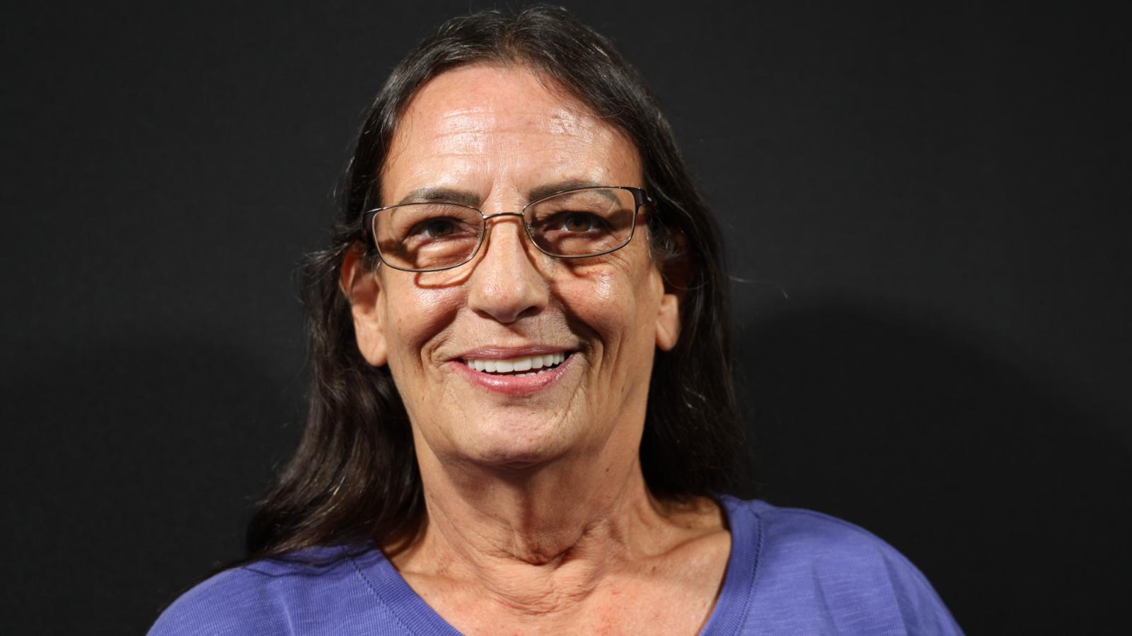 A woman with long dark hair and glasses, wearing a purple shirt, smiles against a black background.