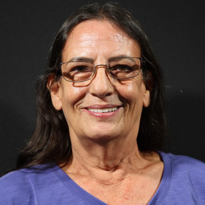 A woman with long dark hair, glasses, and a light purple shirt smiles in front of a dark background.