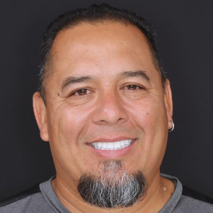 A man with a short, black and gray beard, wearing earrings, smiles in front of a plain black background.