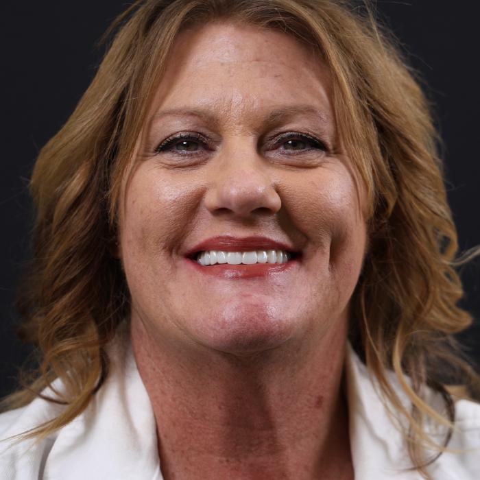 A smiling woman with shoulder-length wavy hair wearing a white shirt is pictured against a dark background.