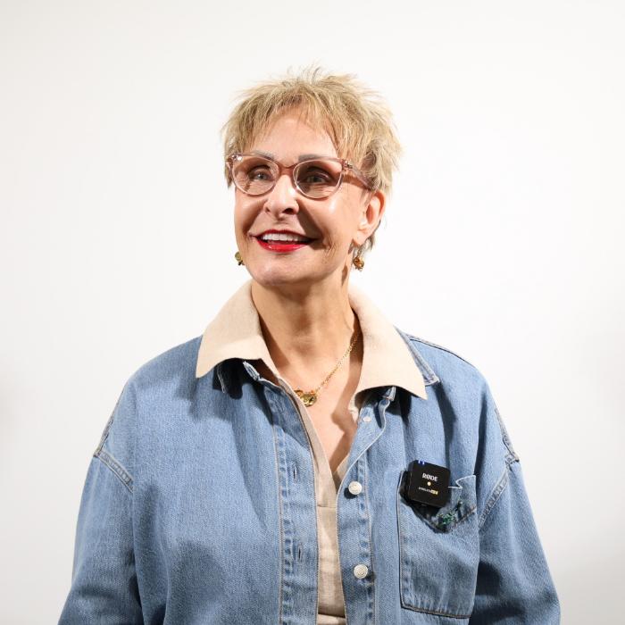 A person with short blonde hair and glasses smiles while looking slightly to the side, dressed in a denim jacket over a beige shirt.