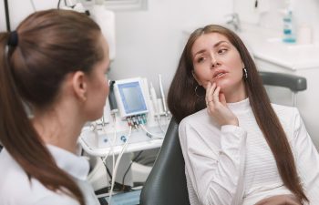 A woman in a dentist's chair touches her face, looking concerned, while a dental professional listens. Dental equipment is visible in the background.